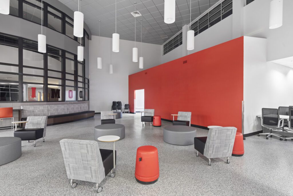 Modern office lounge area with a bold red accent wall, casual seating, media center conversion, and pendant lights.