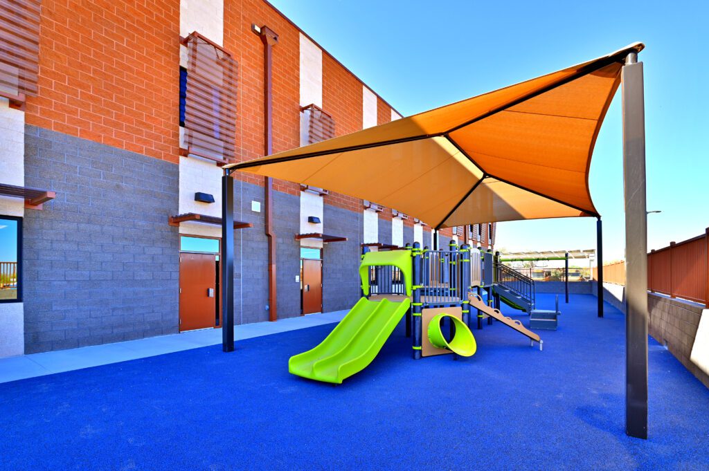 Colorful playground with a shade canopy next to a brick building on the Blackwater Community School campus replacement.